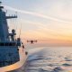 Modern naval vessel patrolling a calm sea at sunset, with a surveillance drone deploying above the ocean surface for a maritime security operation – AI-generated artist’s impression.